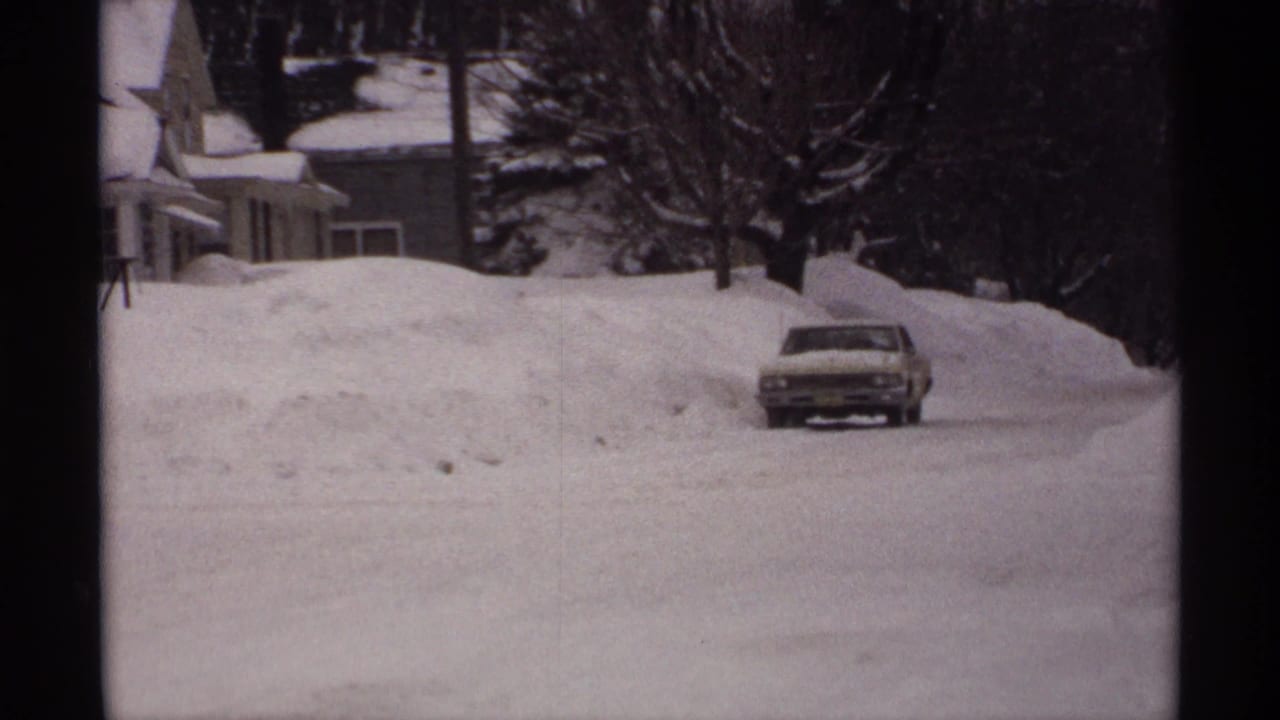 Snow-covered street with a classic car near high snowbanks and houses.