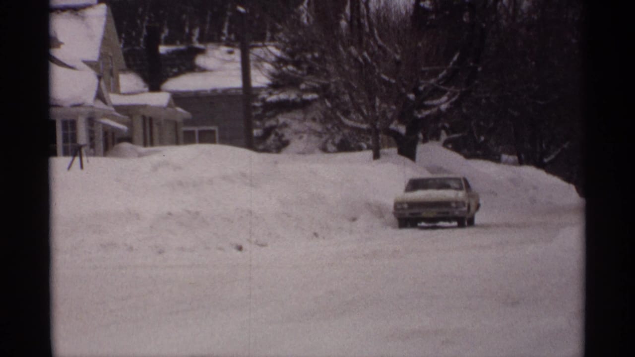 Car on a plowed snowy road beside houses and deep winter snowbanks.