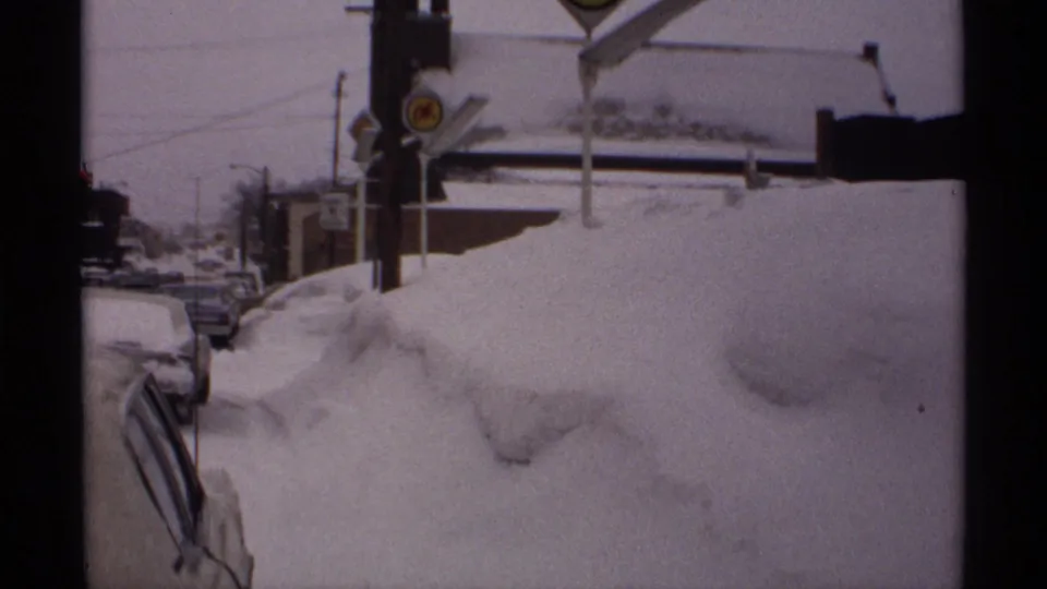 Snowbound Wisconsin street scene with parked cars, deep banks, poles, and a visible crossing sign in 1971 8mm footage.
