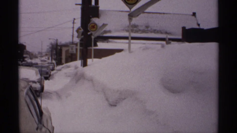 1971 Wisconsin 8mm frame showing deep snowbanks, parked cars partly covered by snow, roadside signs, and utility poles along a winter street.