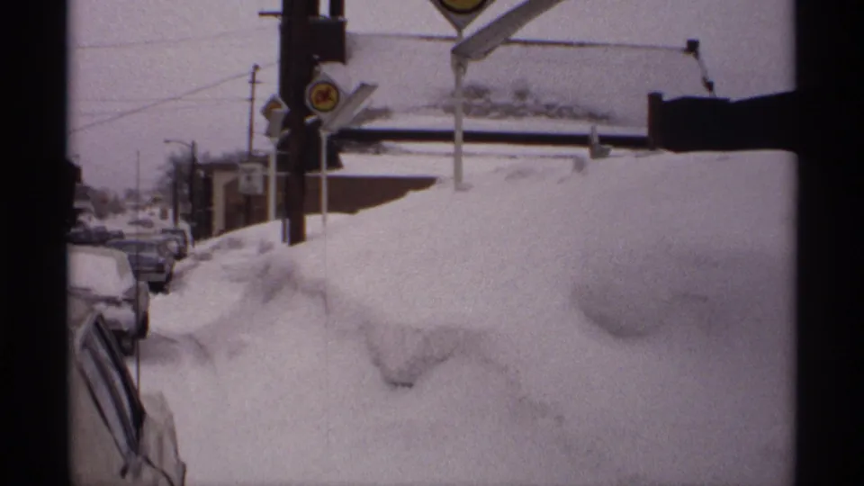 1971 Wisconsin winter street view with a high snowbank, parked cars along the curb, utility poles, overhead wires, and roadside signage.