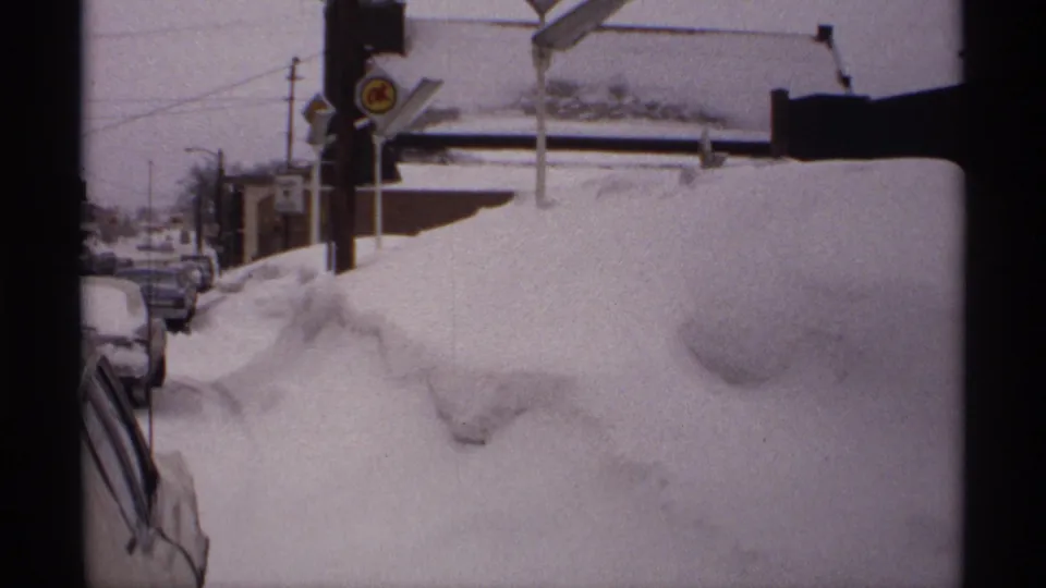 1971 Wisconsin 8mm view of a snow-choked street with cars partly hidden, signage near the sidewalk, utility lines, and a low building beyond.