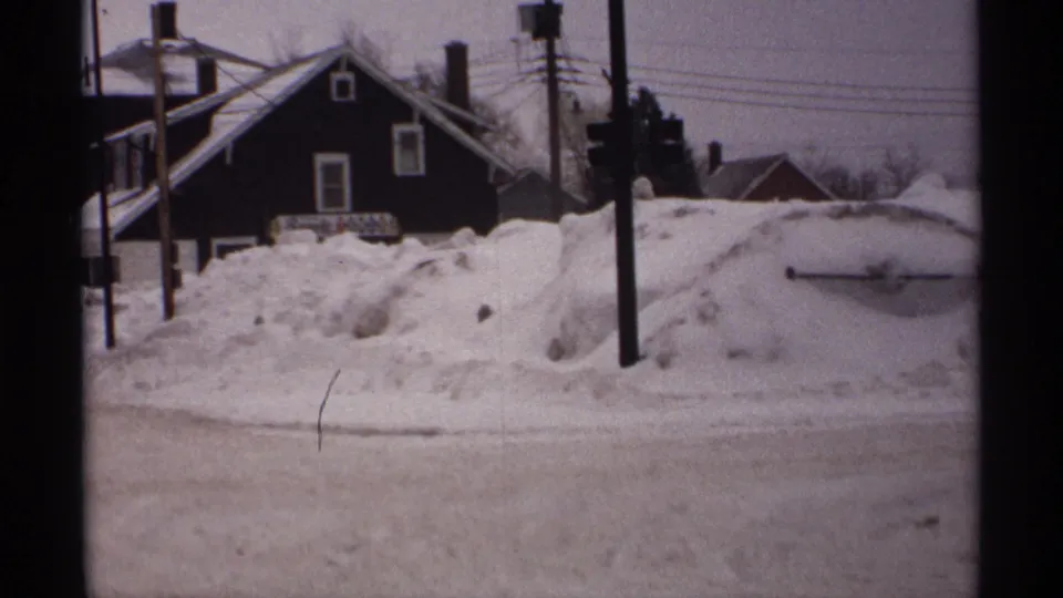 1971 Wisconsin frame showing a dark residential house behind tall snowbanks, utility poles, overhead wires, and a slushy winter roadway.