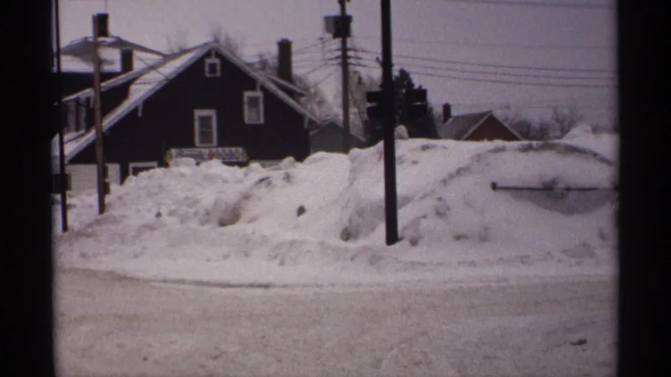 1971 Wisconsin 8mm frame of a snow-covered residential street with deep roadside drifts, a dark pitched-roof house, poles, and wires.