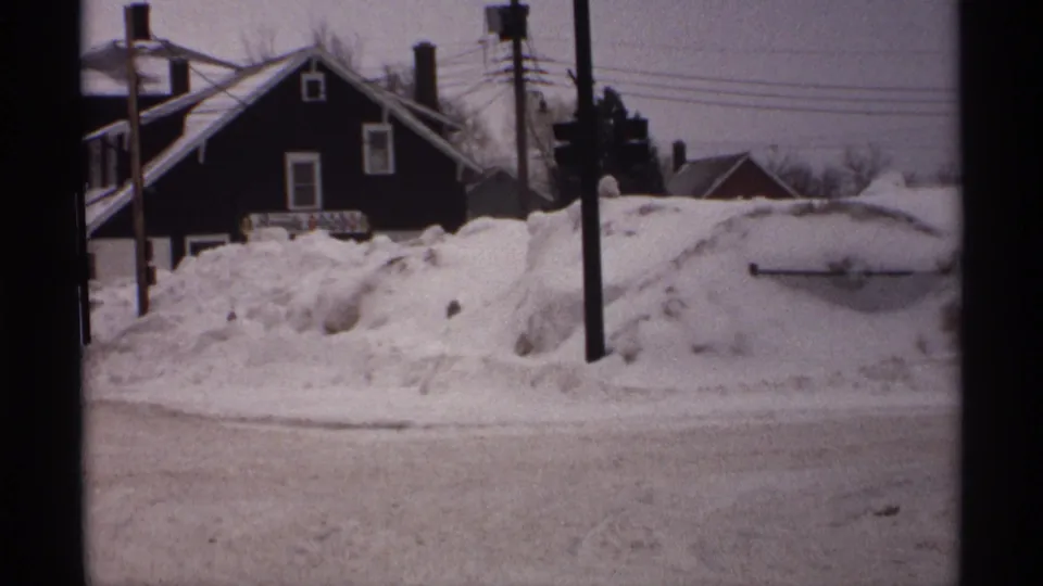 1971 Wisconsin winter frame showing heavy snowdrifts beside a calm street, with houses, bare trees, utility poles, and overhead wires in view.