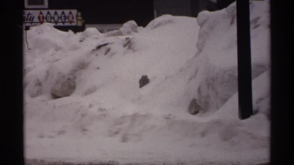 1971 Wisconsin winter frame showing roadside snow piled high, a dark utility pole, building edges, and partially visible commercial lettering.
