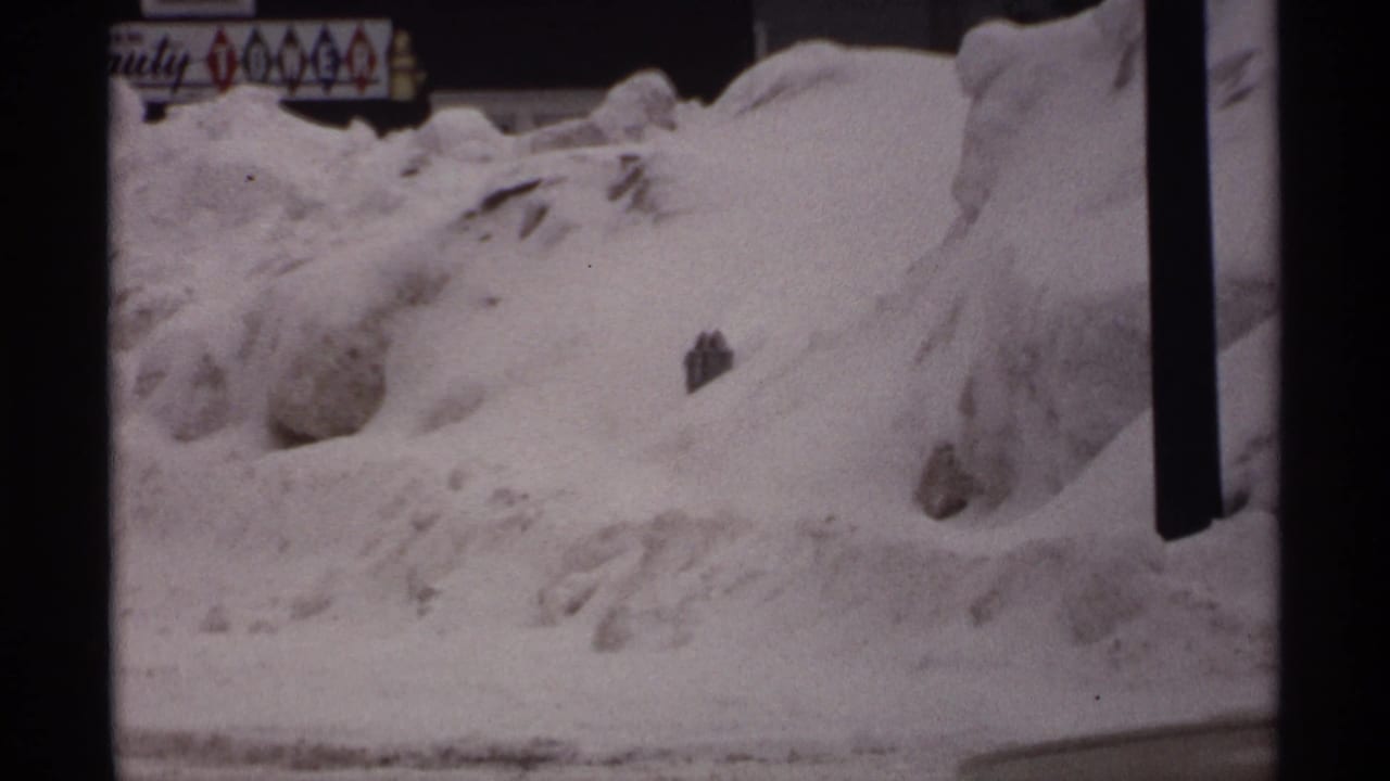 Snowbanks beside a street with a storefront sign, dark posts, and part of a parked or passing car.