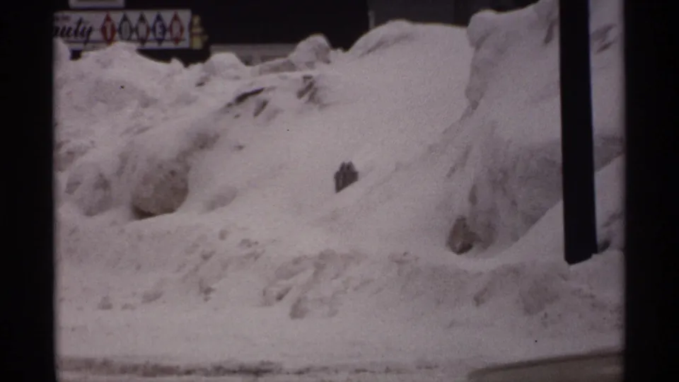 1971 Wisconsin 8mm frame showing deep snowbanks, dark vertical posts, a partial car, and a storefront sign after a winter storm.