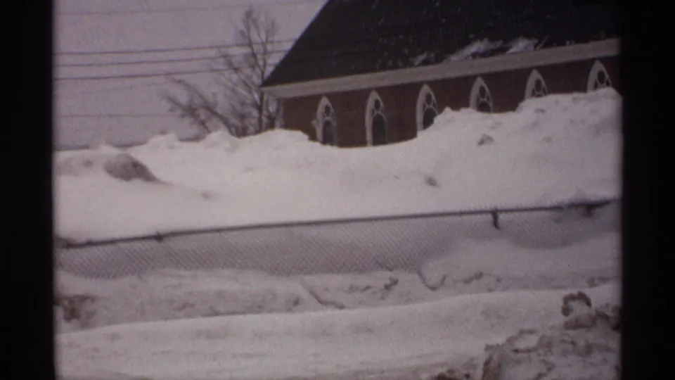 1971 Wisconsin 8mm frame showing a brick church with arched windows behind deep snowbanks, utility wires, and a chain-link fence.