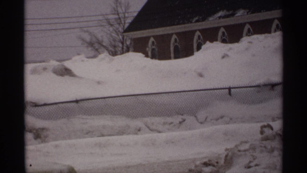 Snow-covered street view with chain-link fence and arched-window brick building beyond.