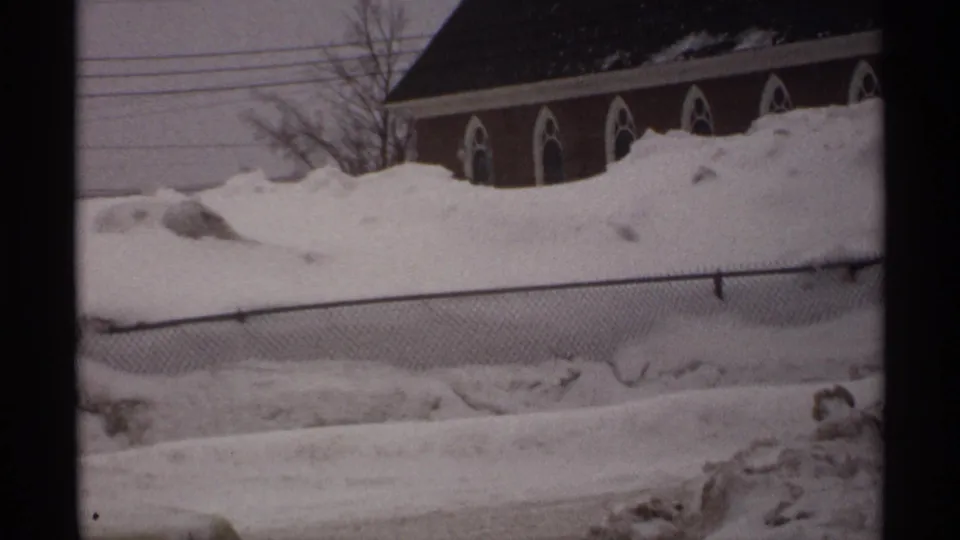 1971 Wisconsin home-movie frame showing deep snow, a chain-link fence, and a brick building with arched windows.