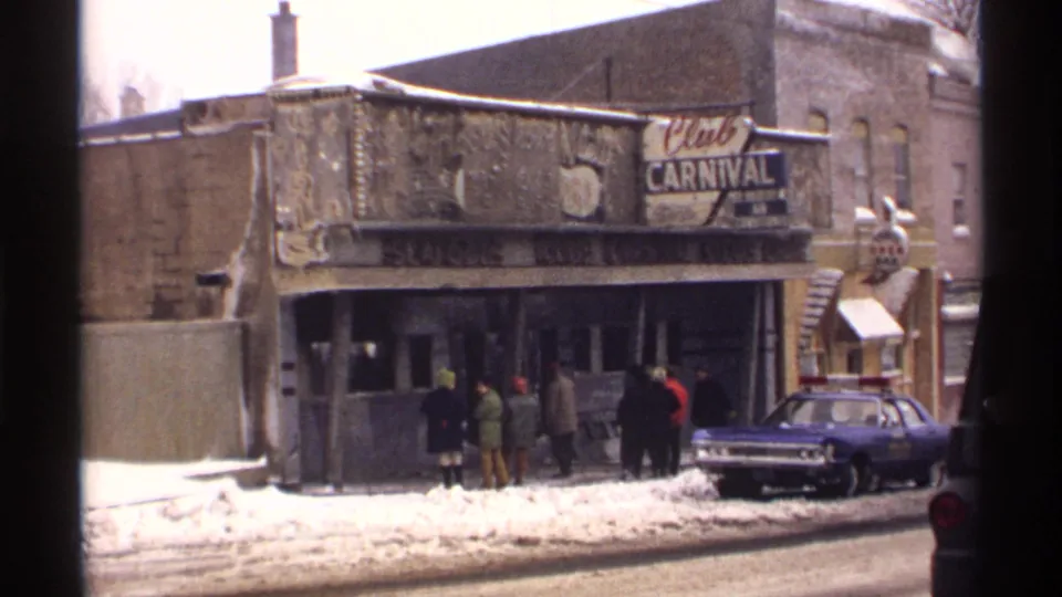 1971 Wisconsin 8mm frame of people waiting near the Club Carnival entrance beside snowbanks and a parked police car.