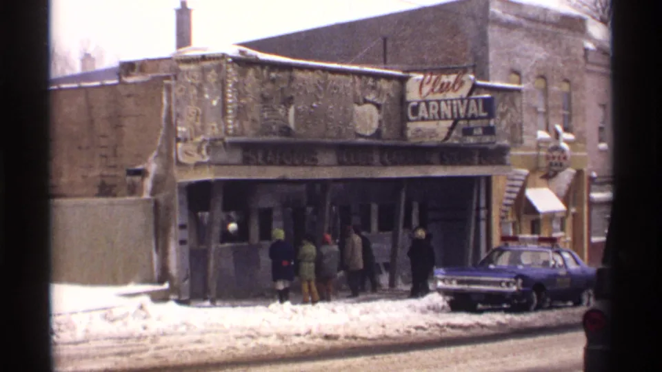 1971 Wisconsin home-movie view of Club Carnival, showing bundled pedestrians, storefront glass, snow, and a police car at the curb.