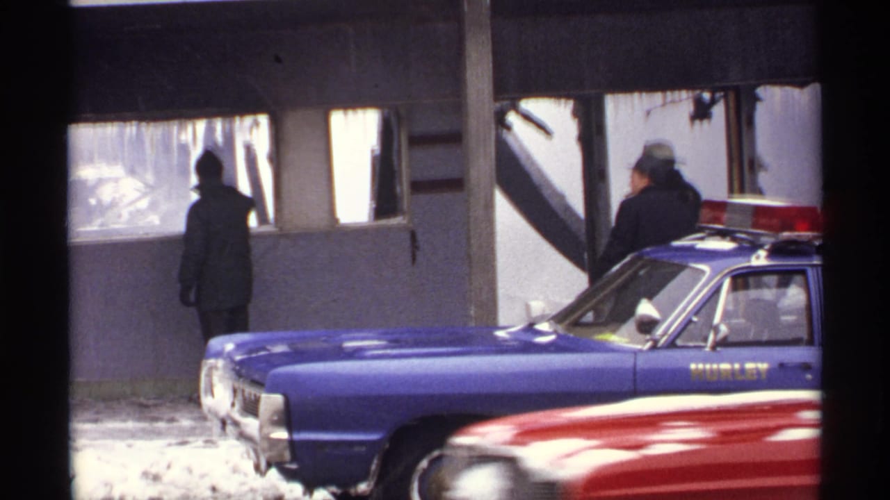 Blue Hurley Police car parked by a damaged snowy building as two bundled adults stand near the entrance.