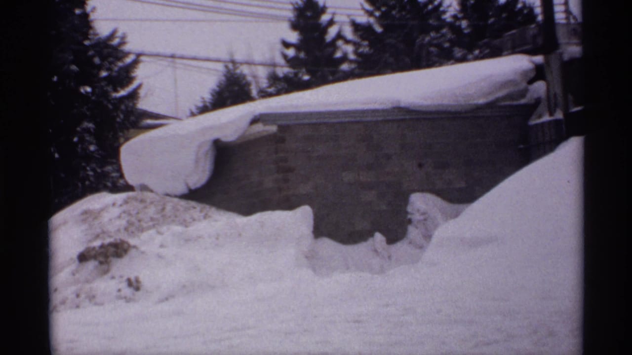 Snowbound Wisconsin scene with a low brick outbuilding partly buried by drifts, evergreen trees behind it, and utility wires overhead.
