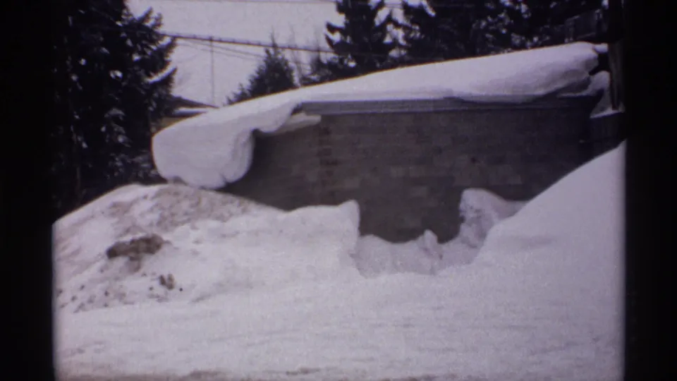 1971 Wisconsin winter frame showing a snow-covered brick structure nearly surrounded by drifts, with dark evergreen trees and utility wires beyond.