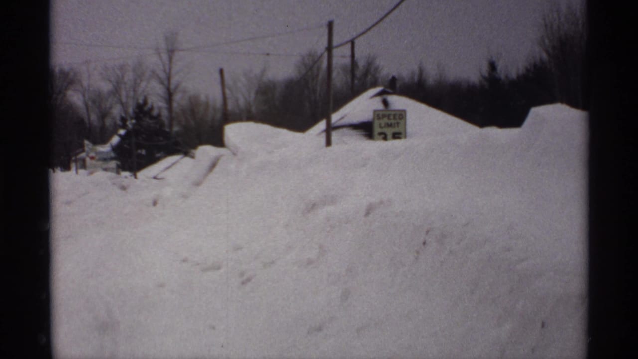 Snow-covered rural road with high snowbanks and a partially buried speed limit sign.
