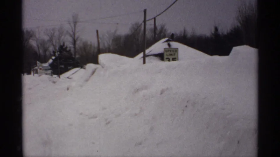 1971 Wisconsin 8mm frame showing a snowy rural road, deep plowed banks, bare trees, utility poles, and a partly buried speed limit sign.