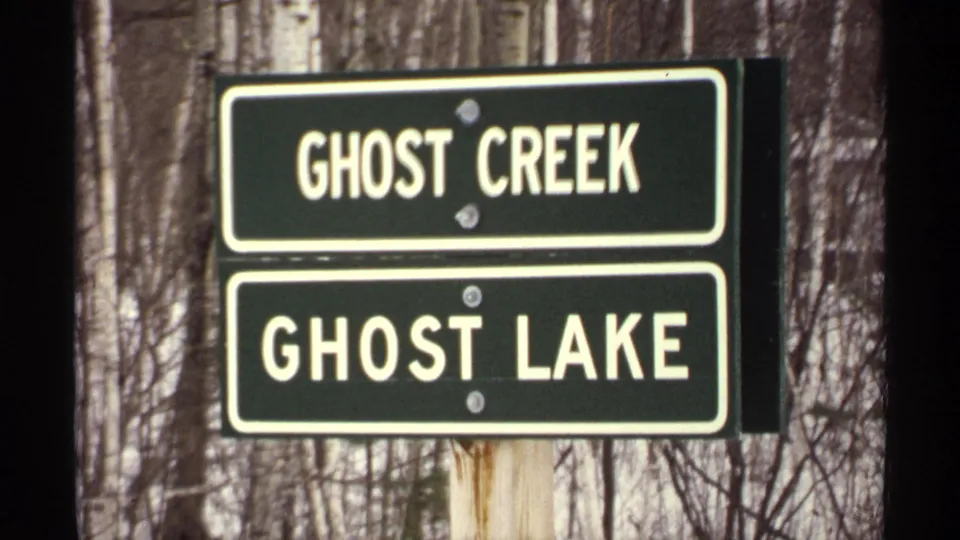1971 Wisconsin 8mm close-up of green Ghost Creek and Ghost Lake signs on a post, with leafless trees and snow behind them.