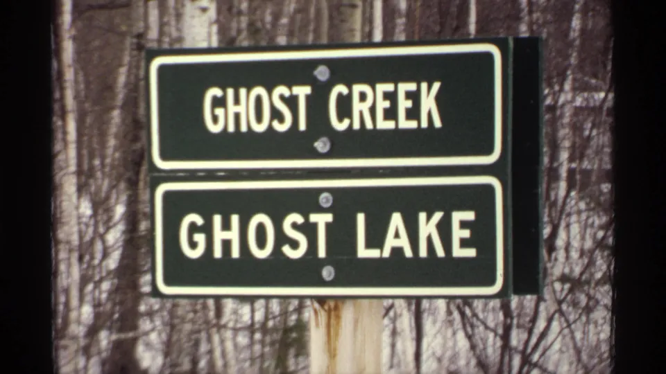 Close 1971 Wisconsin 8mm view of stacked Ghost Creek and Ghost Lake signs, with white lettering, metal fasteners, and winter woods behind.