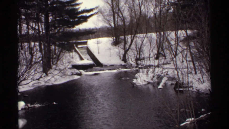 1971 Wisconsin 8mm frame of a dark creek running through snow-dusted woods, with bare branches and a small concrete crossing or structure ahead.