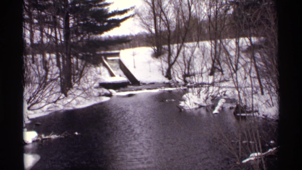 Snow-covered stream and wooded banks with a narrow bridge or crossing in the distance.