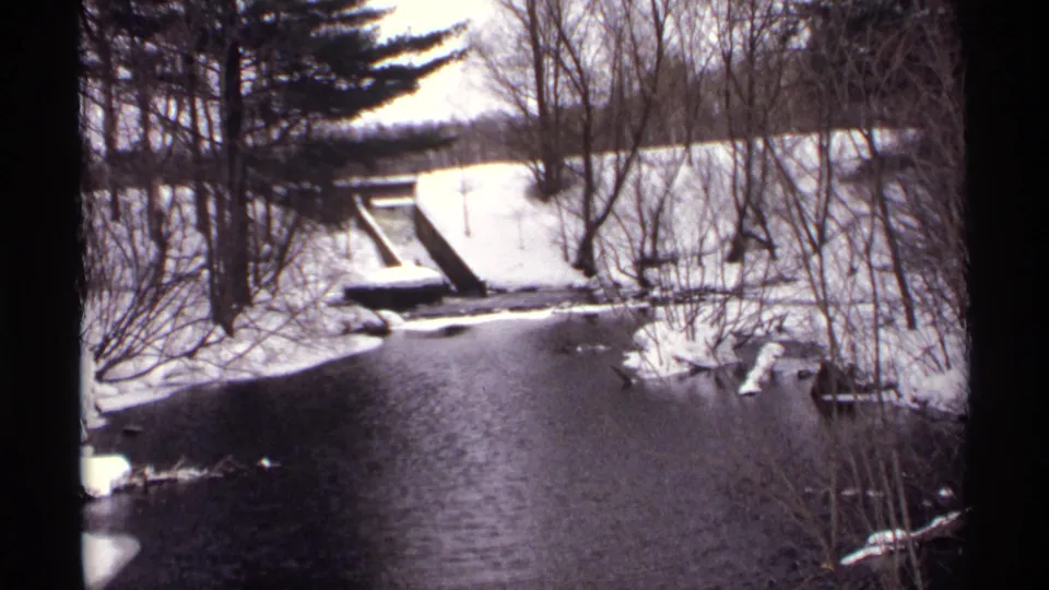 1971 Wisconsin 8mm view of a snowy wooded stream with a narrow bridge or crossing framed by leafless trees and evergreens.