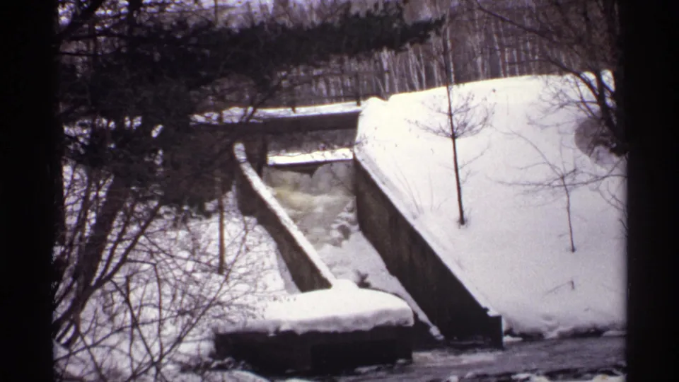 1971 Wisconsin 8mm frame showing a concrete bridge or spillway structure beside snowbanks and an icy stream in wooded winter scenery.