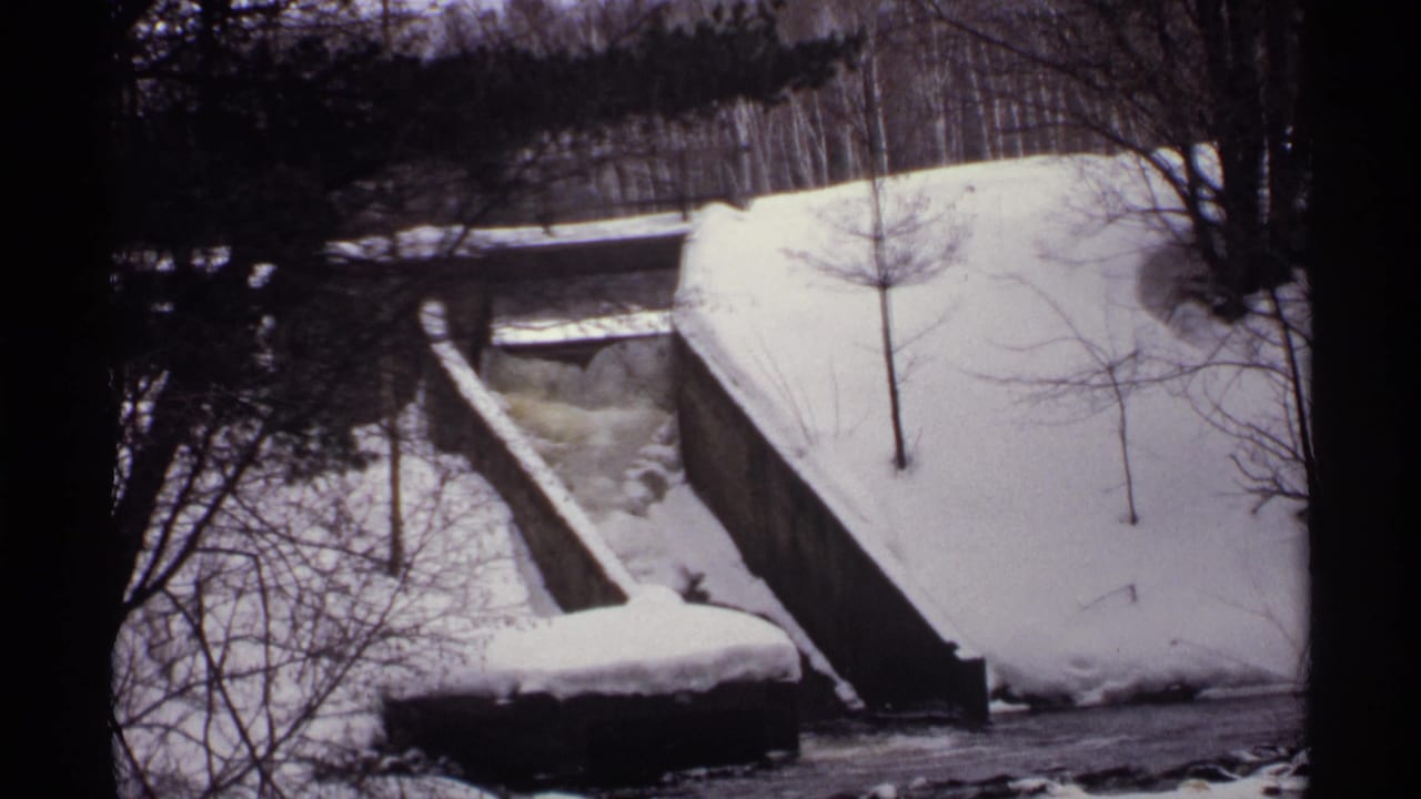 Snow-covered concrete structure beside a dark flowing winter stream in leafless woods.