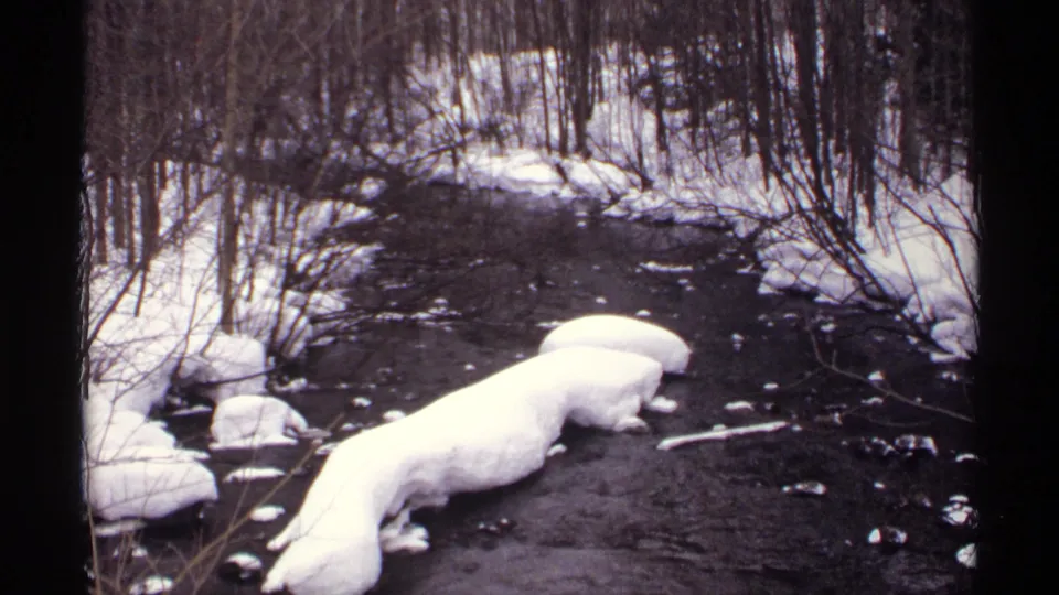 1971 Wisconsin 8mm frame showing dark river water, rounded snow-covered banks, rocks, and dense leafless winter trees.