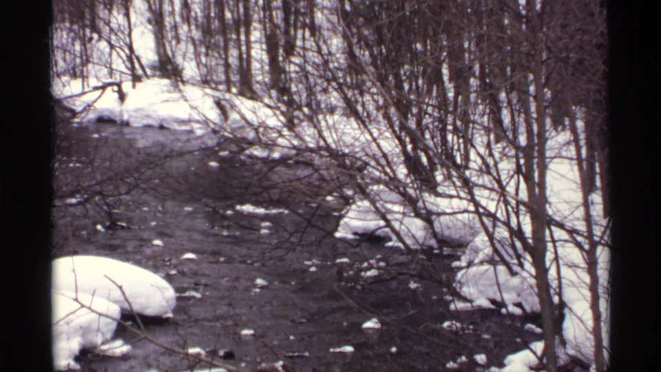 1971 Wisconsin 8mm view down a narrow snowy river corridor with bare brush, rocks, and dark moving water.
