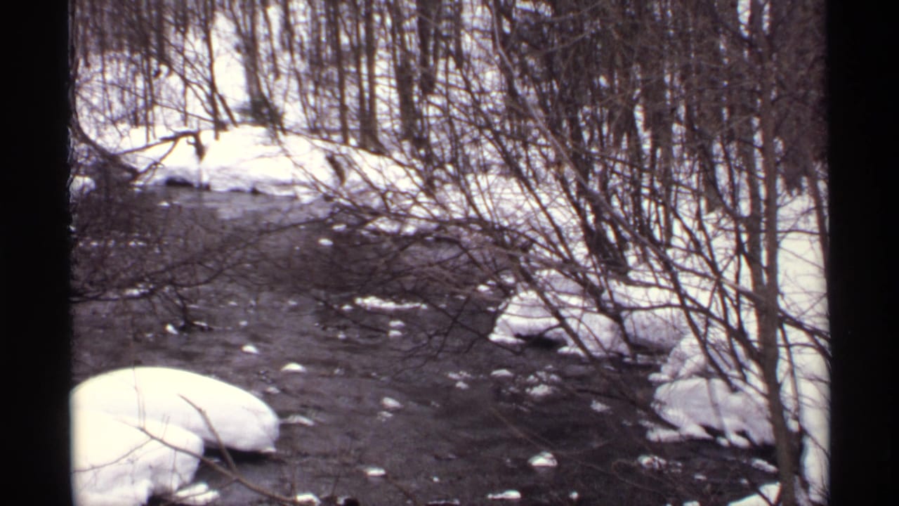 1971 Wisconsin home-movie frame of a snowy wooded river with dark water, ice patches, and bare winter brush.