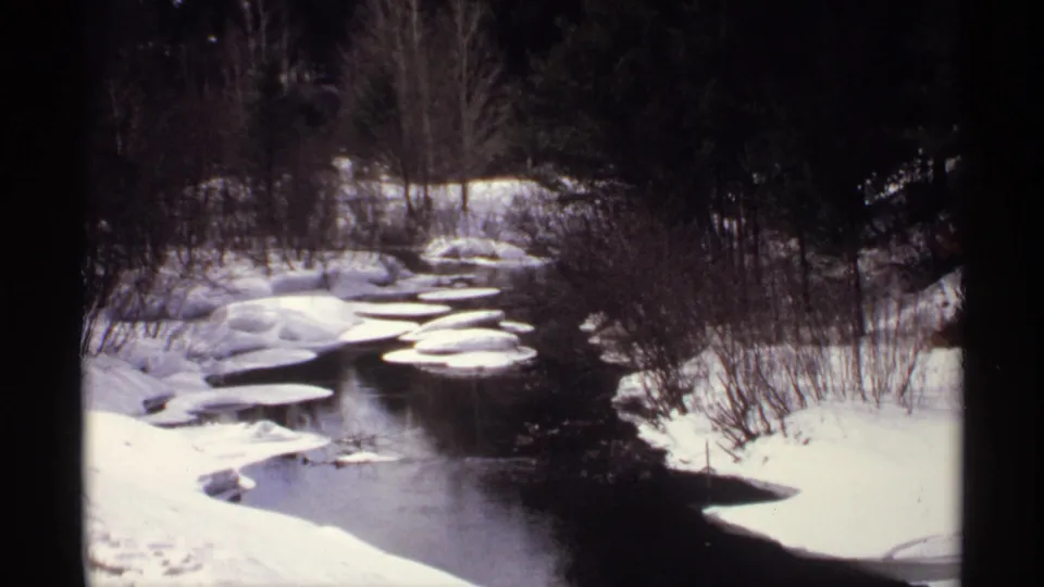 1971 Wisconsin home-movie frame showing a snowy stream bend with dark water, ice floes, and winter brush.