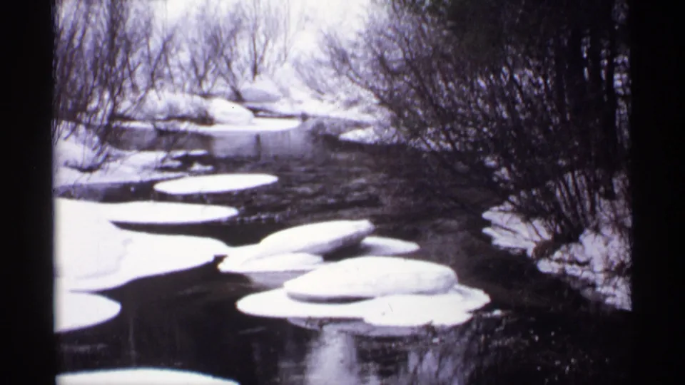 1971 Wisconsin 8mm frame with dark flowing water, bright snow shelves, round ice floes, and winter shrubs.
