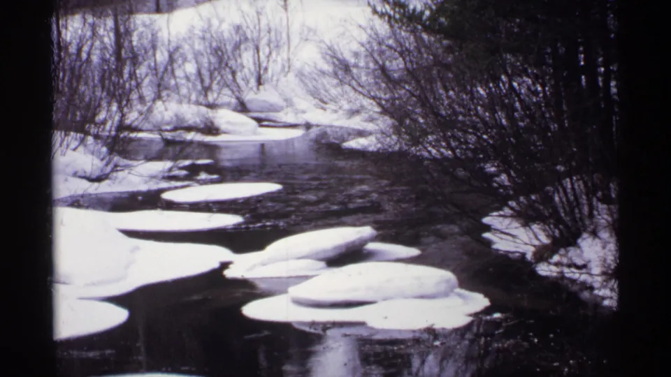 A rural Wisconsin winter river view shows dark current, white ice floes, snowy edges, and leafless trees in 1971 home-movie footage.