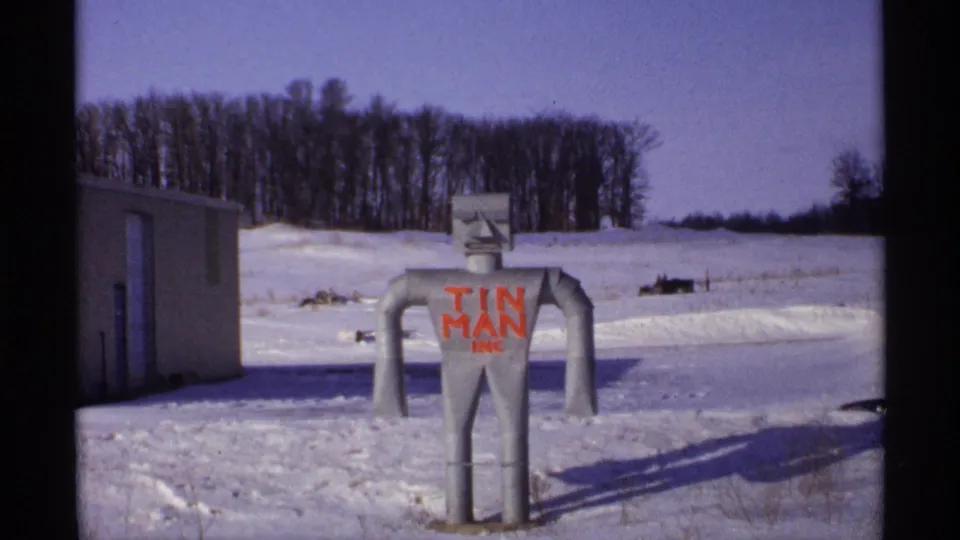 A 1971 Wisconsin winter frame shows a metal tin man figure marked "TIN MAN INC" standing in snow near a small building and bare trees.