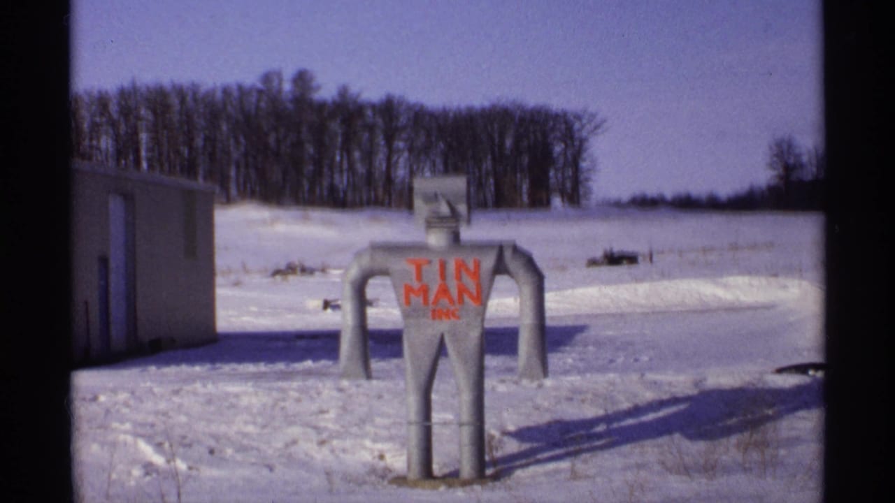 Tin man sculpture standing in snow beside a small building and bare trees.