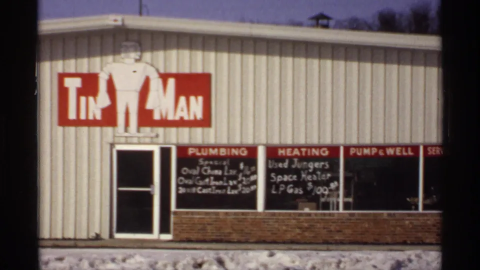 1971 Wisconsin storefront shows a large red-and-white Tin Man sign above windows with chalkboard plumbing, heating, and pump or well notices.