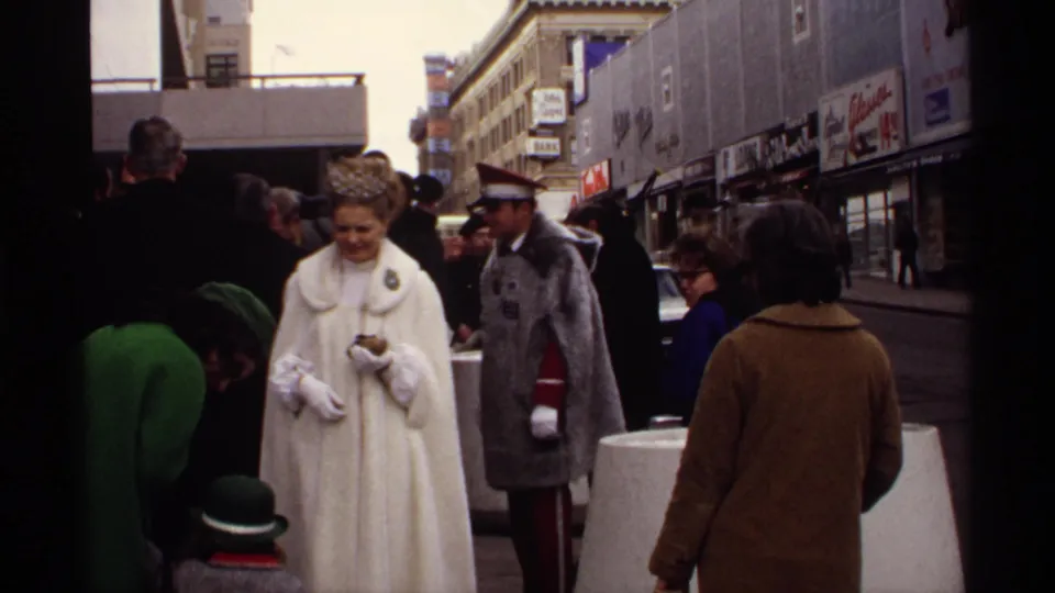 1971 Wisconsin parade scene with onlookers, storefronts, a woman in a white coat and tiara, and a uniformed man.