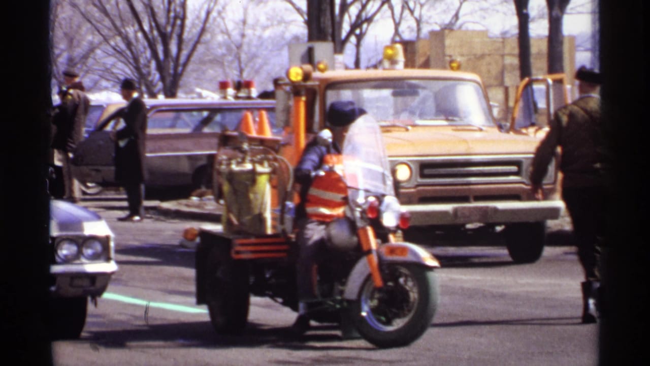 Men stand near a motorcycle with sidecar equipment, yellow truck, and classic cars.