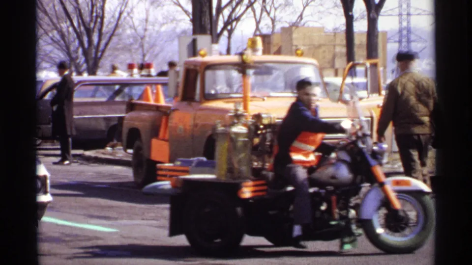 1971 Wisconsin home-movie frame of a vested man riding a motorcycle with sidecar equipment beside a yellow service truck.