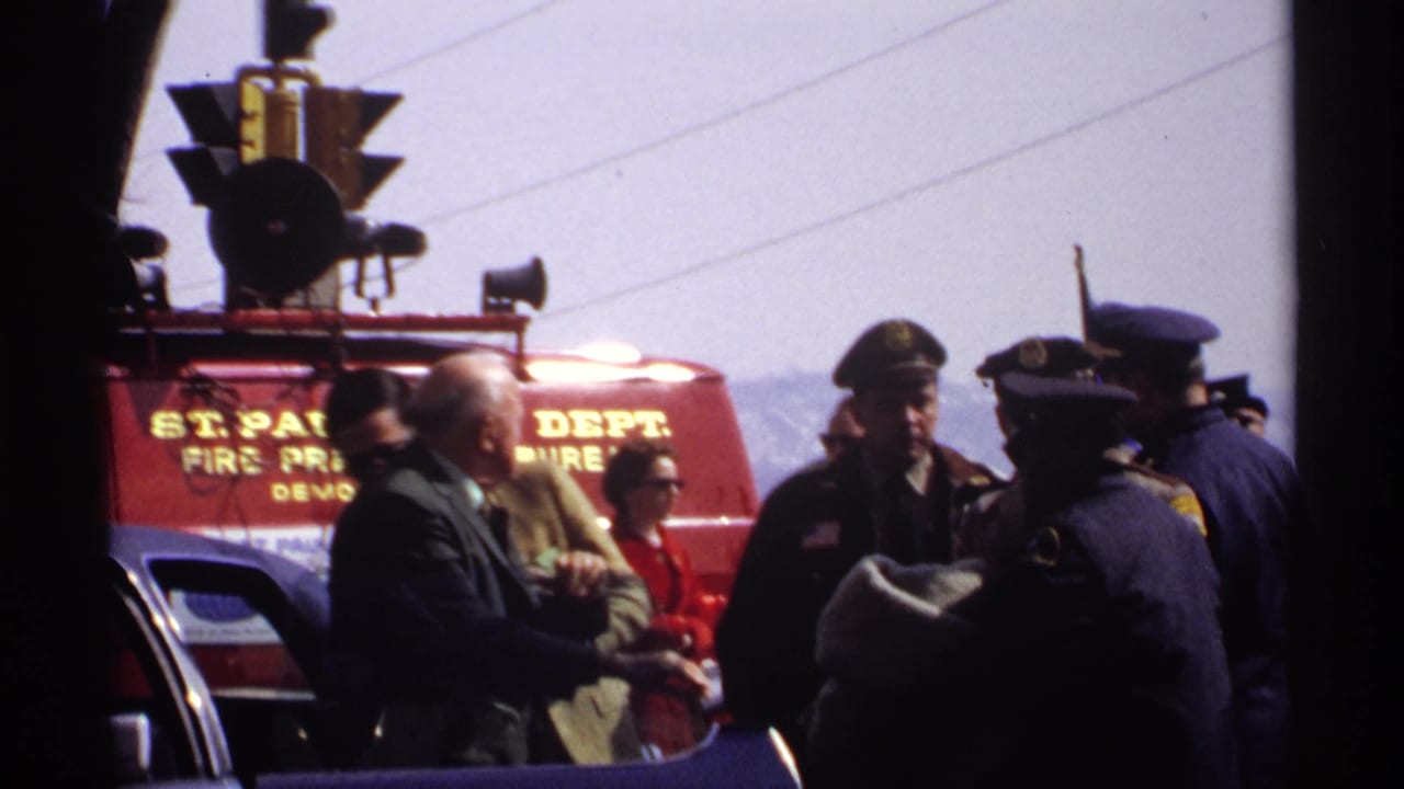 Police officers and suited men remain clustered near a red fire department vehicle, with traffic lights and street details visible in the 1971 parade footage.