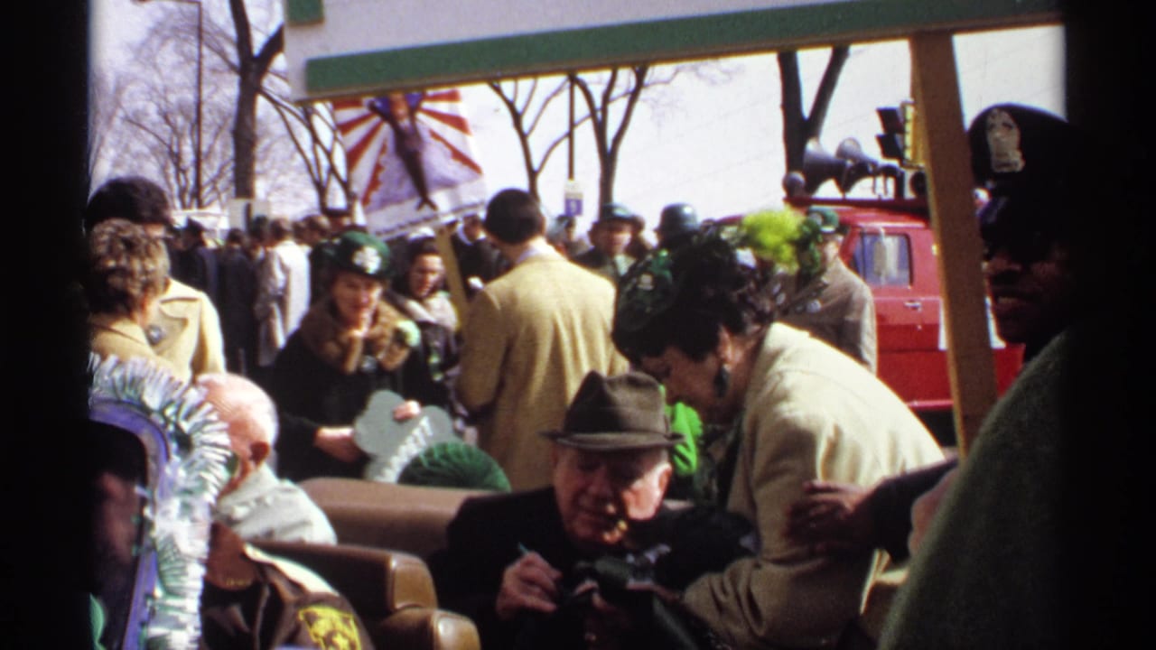 Crowd members in coats and green hats watch the parade from the sidewalk.