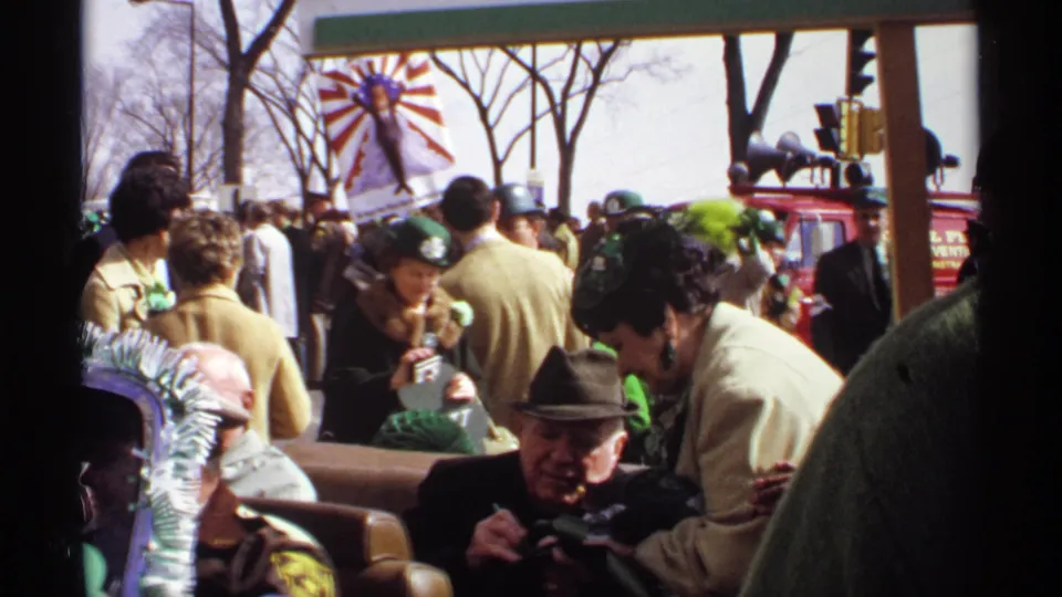 The crowd scene continues with seated and standing spectators, green St. Patrick's Day accessories, a red fire department vehicle in the background, and city street fixtures visible.