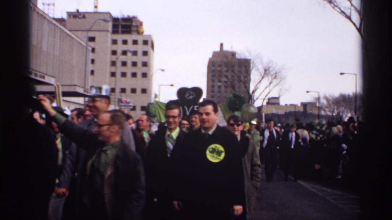 Parade marchers and spectators fill a city street, with men in green accessories and tall buildings behind them.