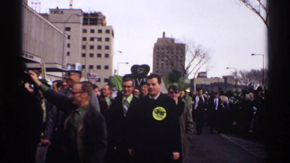1971 Wisconsin parade footage shows people marching past a curbside camera, some wearing green hats and buttons, with urban buildings in the background.