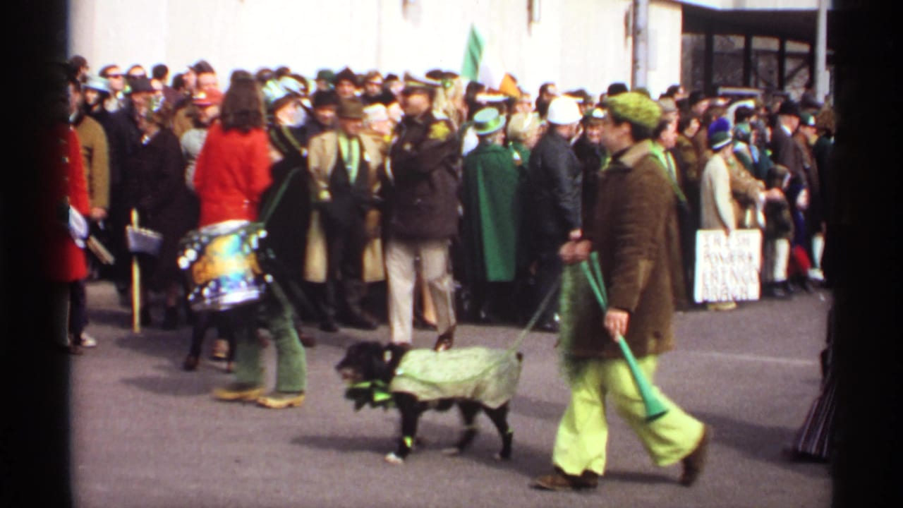 Crowd gathered along a building as a drummer, green-clad marchers, and a costumed dog pass by.