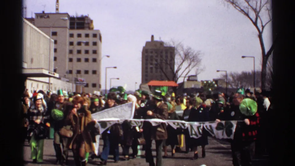 A 1971 Wisconsin St. Patrick's Day parade group advances with a long banner, green hats, and dense urban spectators near buildings including a YWCA sign.
