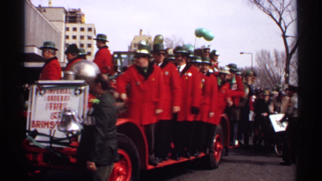 Red-coated parade group riding an antique fire vehicle on a city street.