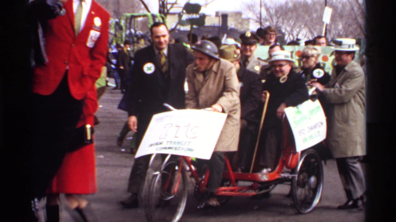 Older participants in a Wisconsin parade ride a red tricycle-style vehicle with hand-lettered signs while spectators and other marchers stand nearby.